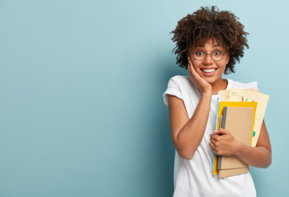 Pleasant looking Afro American woman holds notepads, papers, studies at college, glad to finish studying, keeps hand on cheek, wears white t shirt, isolated on blue background with blank space